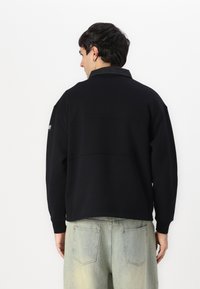 Sudadera negra con un cuello estructurado, puños acanalados y detalles en el panel trasero. Fabricada en un tejido suave; combinada con jeans de colores claros.
