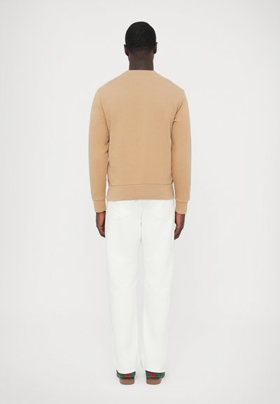 Bež bombažni pulover z okroglim izrezom, rebrastimi manšetami, kombiniran z belimi hlačami z ravnimi hlačnicami in rjavimi čevlji z zelenimi in rdečimi poudarki.