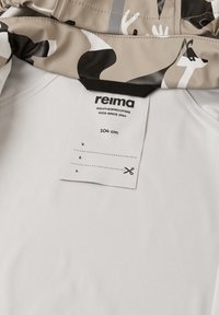 Veste pour enfants Reima avec un motif beige et noir, portant une étiquette indiquant la taille 104 cm et les instructions d'entretien. Texture de tissu lisse.