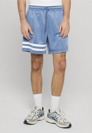 Mann trägt hellblaue Sportshorts mit weißen Streifen am Saum, weiße Kniesocken und silberne Laufschuhe, steht auf grauem Boden.