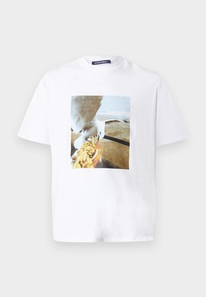 Mees seisab ühevärvilise taustaga, kandes valget graafilist T-särki koos kajaka ja friikartulitega, musti paelaga pükse ja musti tossusid.