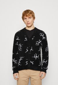 Juodas kardiganas su pūkuota tekstūra, su balta abstrakčia šakų raštu, V formos iškirpte ir sagomis priekyje. Dėvimas ant juodos marškinėlės.