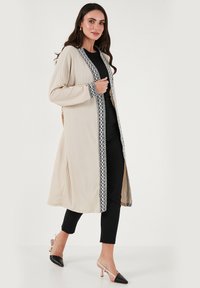 Beige long cardigan with black decorative trim, long sleeves, side slits, ribbed texture, worn over a black outfit and paired with black heels.