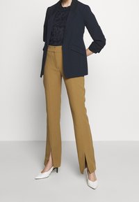 Námořnický blazer se strukturovaným designem, doplněný hořčicovými kalhotami se bočními rozparky, které ladí s bílými špičatými podpatky.