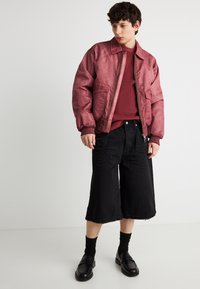 Νέος άνδρας με μπορντό bomber τζάκετ, πουλόβερ πλεκτό στο ίδιο χρώμα, φαρδιά μαύρη cropped παντελόνια, μαύρες κάλτσες και μαύρα loafers.