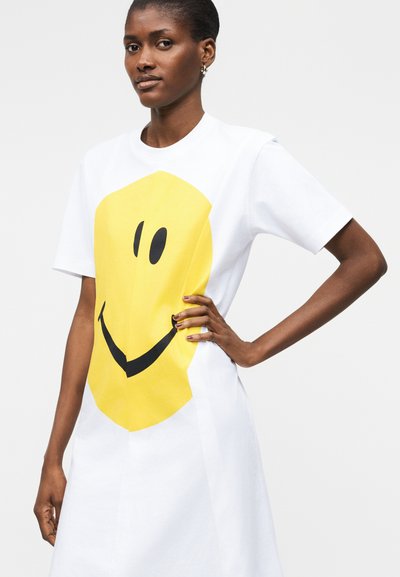 Femme portant une robe blanche à manches courtes avec un grand motif visage souriant jaune, debout avec une main sur la hanche devant un fond uni.