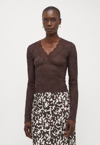 Brown lace long-sleeve top with a scalloped neckline, featuring intricate floral patterns. Paired with a patterned brown and white skirt.