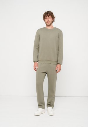 Homme debout devant un fond blanc, portant un sweat-shirt et un pantalon vert olive assortis avec des baskets blanches.