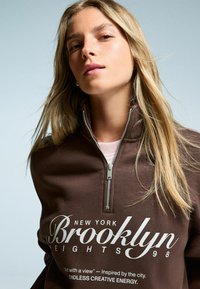 Bruine rits-trui met hoge kraag, met witte tekst "Brooklyn Heights 98" en decoratieve elementen. Gladde textuur.