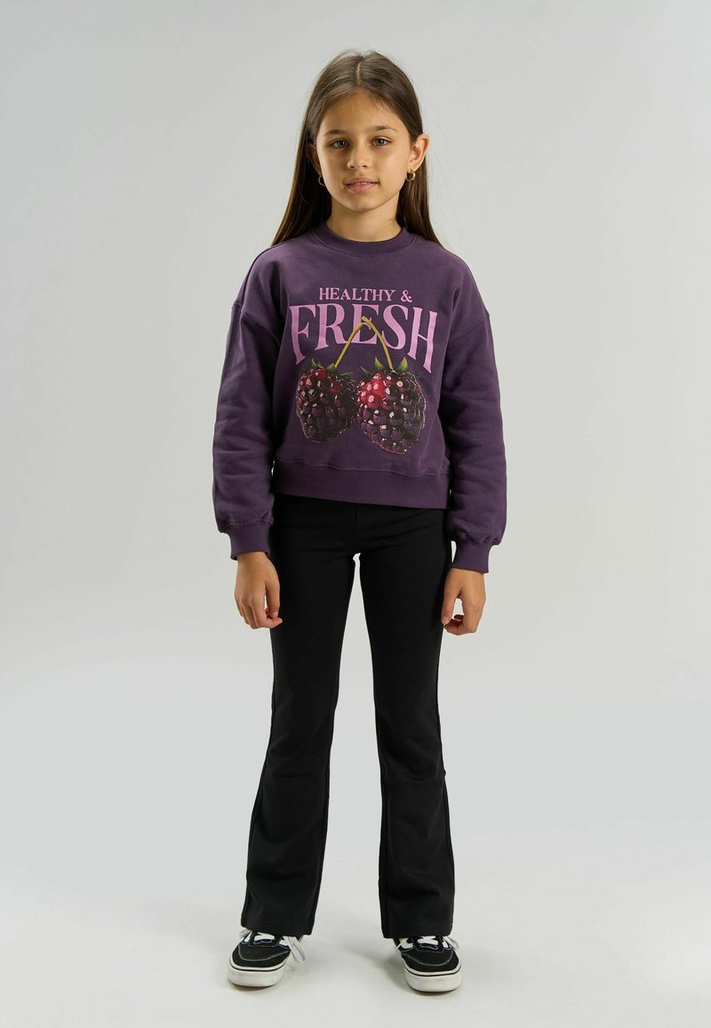 Fille aux longs cheveux bruns porte un sweat-shirt violet avec la mention « Healthy & Fresh » et de grosses cerises, un pantalon noir et des baskets noires avec des semelles blanches.