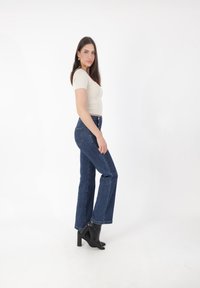 Młoda kobieta stojąca bokiem, ubrana w beżową bluzkę z krótkim rękawem, ciemnoniebieskie dżinsy i czarne botki na obcasie, na tle białego tła.