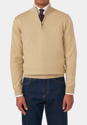Homme portant un pull beige à zip court sur une chemise blanche avec une cravate rayée bordeaux, associé à un jean bleu foncé, sur un fond uni.