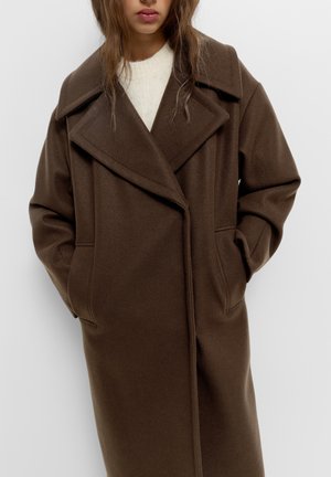 Femme portant un manteau en laine marron oversize avec de larges revers et les mains dans les poches, par-dessus un pull en maille blanc, sur un fond uni.