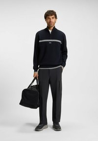 Navy zip-neck pullover with white striped accents, paired with black pants featuring pockets, and a black duffel bag. Gray sneakers complete the look.