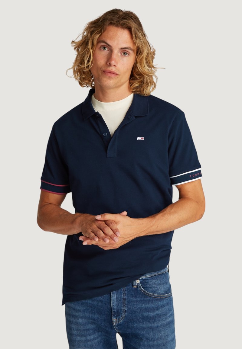 Polo azul marino de algodón, con un cuello corto, botones y puños a rayas en contraste con detalles de marca en las mangas.