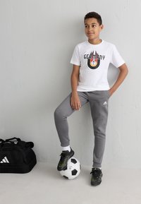 Camiseta blanca con un gráfico de "ALEMANIA", pantalones de chándal grises, zapatos negros con detalles en verde, de pie sobre un balón de fútbol, bolsa de deporte cerca.