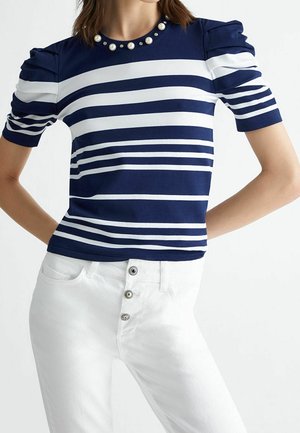 Donna che indossa un top a maniche corte a righe blu navy e bianco con dettaglio colletto di perle e pantaloni bianchi a vita alta con bottoni a vista.