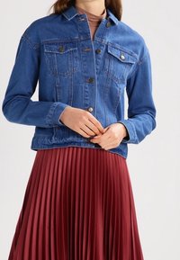 Giacca di jeans di blu medio, con chiusura a bottoni, tasche sul petto e un taglio dritto. Indossata sopra una gonna plissè borgogna.