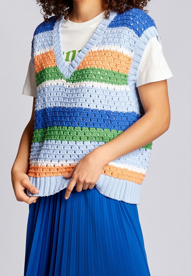 Personne portant un gilet tricoté multicolore avec des rayures bleues, vertes, orange et blanches sur un t-shirt blanc et une jupe plissée bleue.