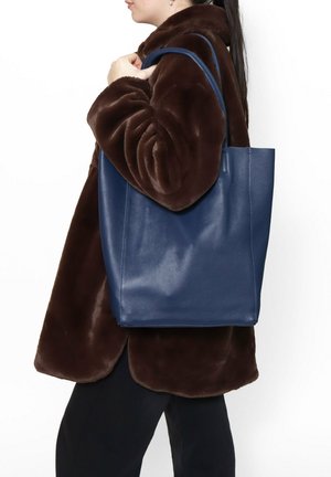 Marineblauwe leren totebag met een gladde textuur, rechthoekige vorm en lange, gebogen hengsels, gedragen naast een bruine bontjas.