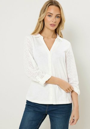 Blonde Frau, die eine weiße Bluse mit Knöpfen und Ärmeln aus Lochspitze sowie dunkelblaue Jeans trägt, vor einem schlichten hellen Hintergrund stehend.