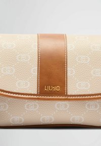 Kabelka z textúrovaného béžového materiálu s bielou logom. Obsahuje hladký hnedý kožený akcent a zlatý logotyp.