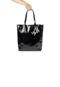 Zwarte glanzende tote bag met twee lange handvatten, met een strak rechthoekig ontwerp en een metalen bedel met de gravure "MH."