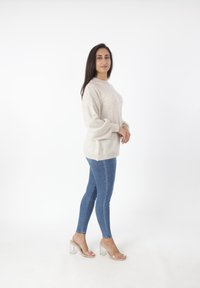 Beige oversized Pullover mit langen Ärmeln, blauen Skinny Jeans und transparenten Blockabsatzsandalen, die vor einem schlichten weißen Hintergrund steht.