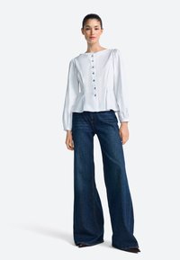 Balta blūze ar augstu kakla izgriezumu un garām pūdera piedurknēm, kurās ir piecas pogas. Valkāta ar plašām tumša džinsa biksēm.