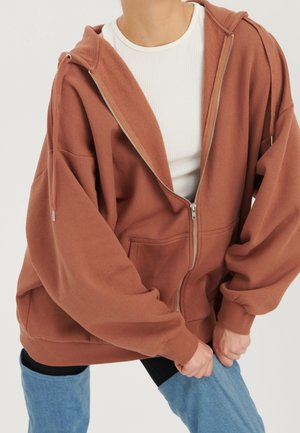 Personne portant un sweat à capuche marron oversize zippé sur un t-shirt blanc côtelé, remontant des leggings bleu clair style denim sur des collants noirs.