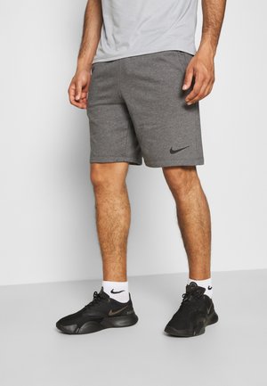 Uomo con pantaloncini grigi Nike, calzini bianchi Nike e scarpe da ginnastica nere Nike che sta in piedi su un pavimento grigio contro una parete bianca.