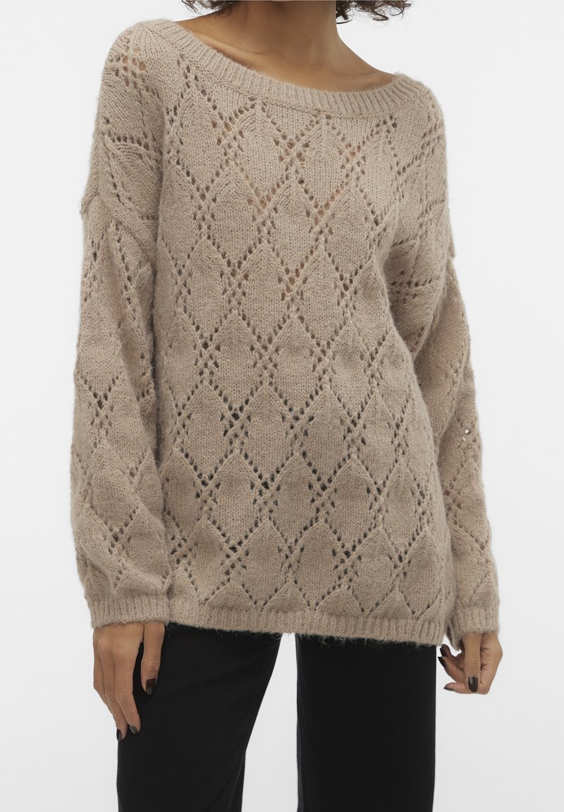Femme portant un pull en maille beige avec un motif ajouré en forme de losange et un pantalon noir sur un fond uni clair.