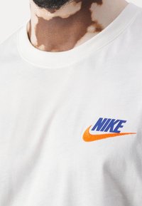 Vit bomullströja med en blå "NIKE"-logotyp och en orange swoosh. Ribbade krage, slät textur och avslappnad passform.