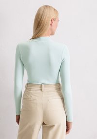 Langärmliges, mintgrünes Oberteil aus dehnbarem Stoff, das in hoch taillierte, beigefarbene Cordhosen mit vertikalen Streifen und Gesäßtaschen gesteckt ist.