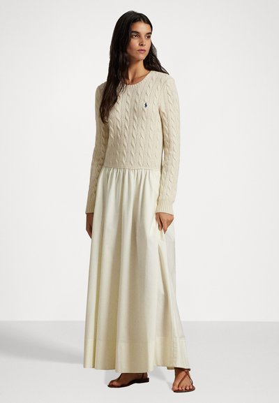 Uma mulher com cabelos longos e escuros veste um vestido de suéter beje com padrão de tramas e uma saia texturizada, usando sandálias rasas castanhas, de pé contra um fundo liso.