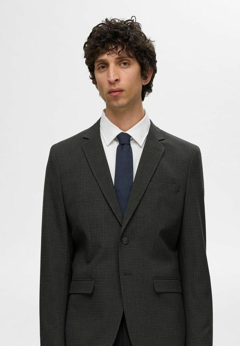 Mørkegrå rutete blazer med klippede lapeller, enkel knappelukking og brystlomme. Kombinert med en hvit skjorte og marineblå slips.