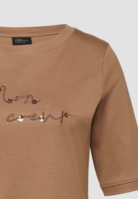 Chemise marron à manches courtes avec texte cursif brodé et petits sequins sur le devant, dotée d'un col rond et d'une étiquette de marque visible à l'intérieur.