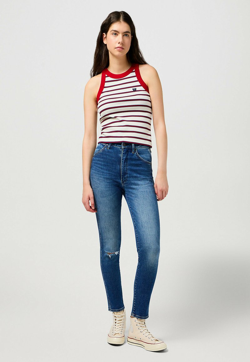 Striped tank top with red trim, paired with fitted blue jeans featuring a distressed knee. Model stands in casual canvas sneakers.