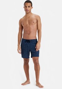 Tamsiai mėlynos spalvos plaukimo šortai su taškuotu raštu, elastine juosmens juosta ir raišteliu. Audinio tekstūra atrodo lygi ir lengva.