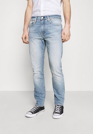 Homme portant un jean slim bleu clair et des baskets montantes noires, debout devant un fond blanc uni.