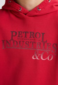 Felpa rossa con una texture morbida, caratterizzata dalla stampa "PETROL INDUSTRIES" in grigio scuro, con dettagli argentati e occhielli per il cordino.