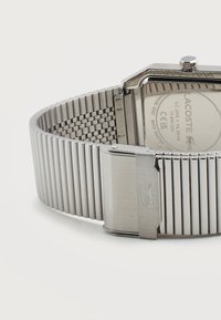 Ιμάντας ρολογιού από ανοξείδωτο ατσάλι σε ασημί χρώμα με κούμπωμα και ορατό χαραγμένο λογότυπο Lacoste στην πίσω πλευρά της κάσας και στο κούμπωμα.