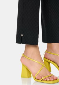 Zwarte geklede broek met een patroon, voorzien van een zilveren accent aan de zoom, gecombineerd met gele sandalen met blokhak en bandjes.