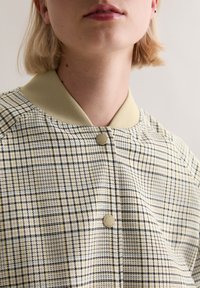 Καρό μπουφάν bomber σε κρεμ και ανοιχτό πράσινο, με ριμπ γιακά, κλείσιμο με κουμπιά πατιλέτα και υφασμάτινη υφή πλεγμένη.