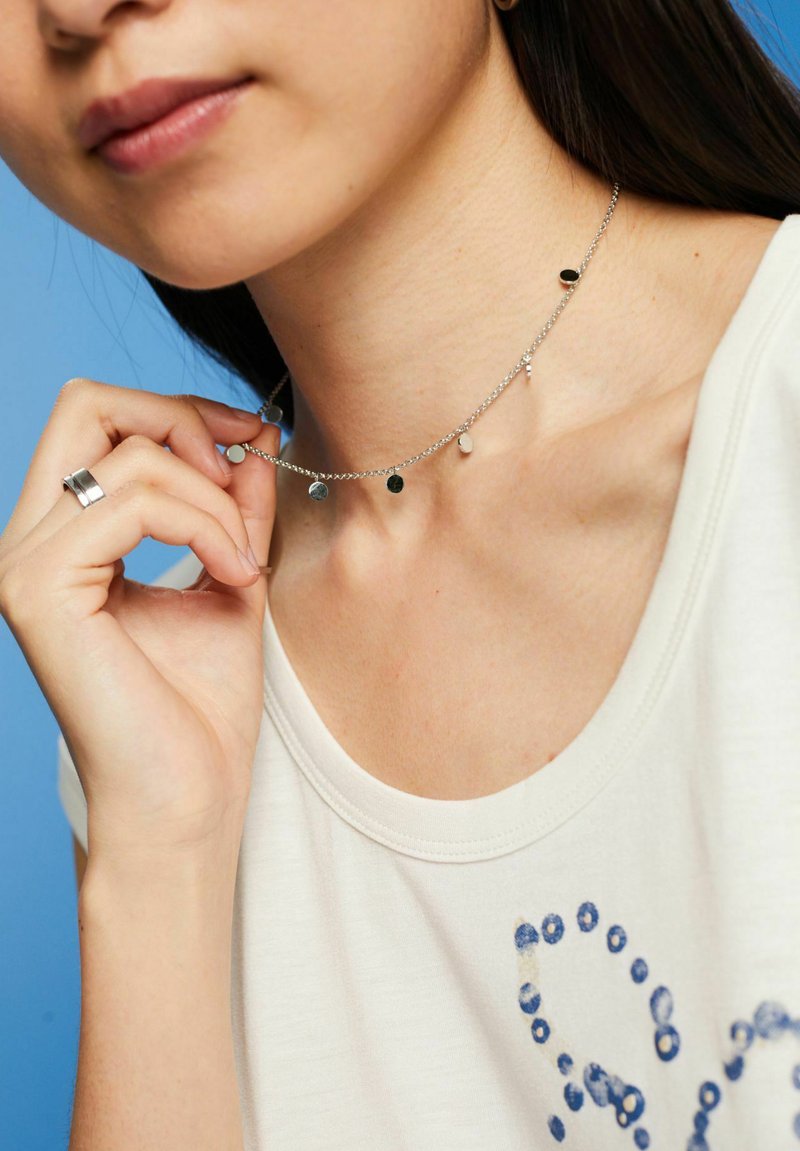 Collana in argento con molteplici pendenti rotondi in finiture nere, bianche e metalliche, indossata con una maglietta chiara.