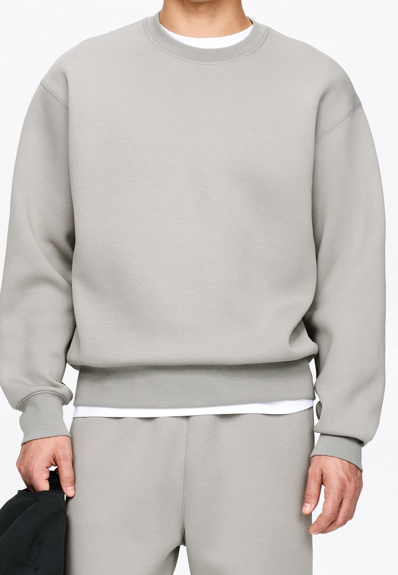 Homme portant un sweat-shirt gris clair à col rond et un pantalon assorti, tenant une casquette noire dans sa main gauche.