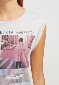 Gray sleeveless t-shirt featuring a printed graphic of a person in a pink coat, with text including "MANIFESTO" and "LIVE FREE AND DREAM."