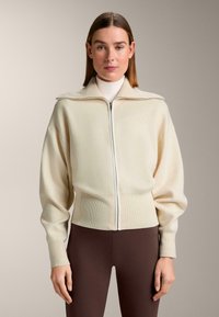 Crèmekleurige zip-up trui met een hoge kraag, geribbelde zoom en manchetten, met een zachte textuur en een relaxte pasvorm. Draagt bijbehorende donkerbruine legging.