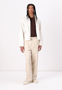 Light beige zip-up jacket, brown t-shirt underneath, loose cream trousers, and light shoes; simple design, relaxed fit, and smooth texture.