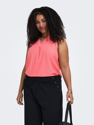 Femme aux cheveux bouclés portant un haut sans manches corail rose et un pantalon noir avec des détails à boutons, tenant un sac à main noir dans sa main droite.